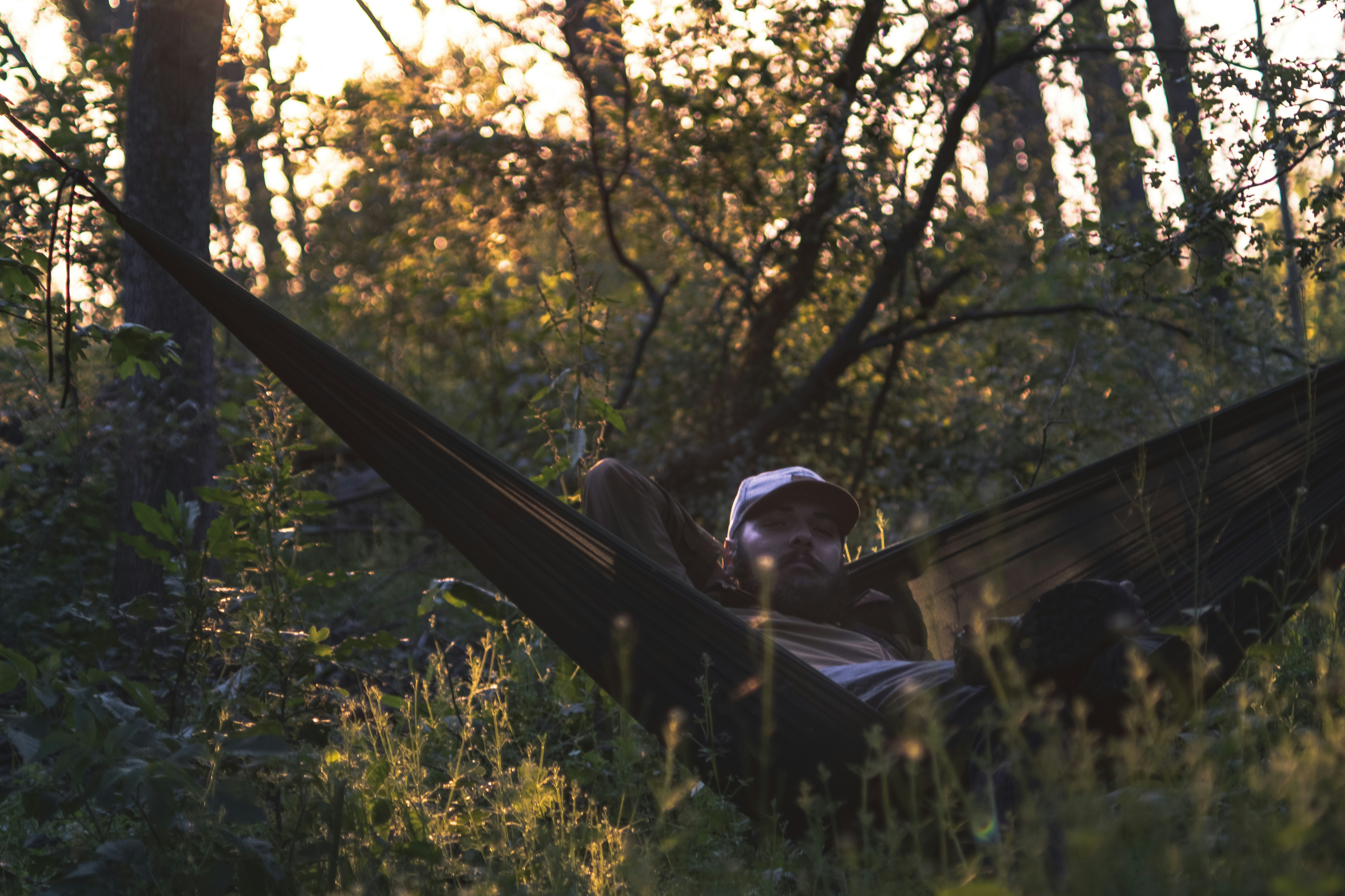 man lying on black hammock surrounded with tall and green trees