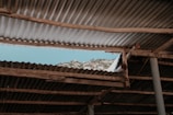A section of corrugated metal roofing with wooden supports reveals a view of a mountainous landscape beneath a clear blue sky. The roofing is aged and weathered, adding a rustic feel to the scene.