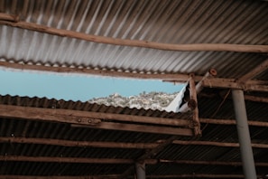A section of corrugated metal roofing with wooden supports reveals a view of a mountainous landscape beneath a clear blue sky. The roofing is aged and weathered, adding a rustic feel to the scene.