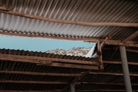 A section of corrugated metal roofing with wooden supports reveals a view of a mountainous landscape beneath a clear blue sky. The roofing is aged and weathered, adding a rustic feel to the scene.