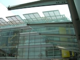 Modern architectural structure with glass and metal elements. The design features overlapping glass panels and metal beams, with reflections and text visible on the glass facade. The lower part of a building is visible with signage indicating shops or businesses like Ozoc and IFC.