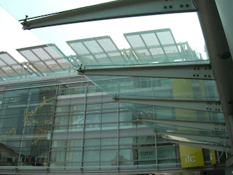 Modern architectural structure with glass and metal elements. The design features overlapping glass panels and metal beams, with reflections and text visible on the glass facade. The lower part of a building is visible with signage indicating shops or businesses like Ozoc and IFC.