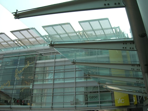 Modern architectural structure with glass and metal elements. The design features overlapping glass panels and metal beams, with reflections and text visible on the glass facade. The lower part of a building is visible with signage indicating shops or businesses like Ozoc and IFC.