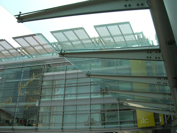 Modern architectural structure with glass and metal elements. The design features overlapping glass panels and metal beams, with reflections and text visible on the glass facade. The lower part of a building is visible with signage indicating shops or businesses like Ozoc and IFC.