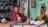 A man and a woman are seated at a cluttered workbench, surrounded by various tools and objects. The man is wearing glasses and a red shirt, while the woman is dressed in a yellow patterned sari. Shelves filled with miscellaneous items are visible in the background, along with a green painted wall adorned with religious images.