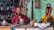 A man and a woman are seated at a cluttered workbench, surrounded by various tools and objects. The man is wearing glasses and a red shirt, while the woman is dressed in a yellow patterned sari. Shelves filled with miscellaneous items are visible in the background, along with a green painted wall adorned with religious images.