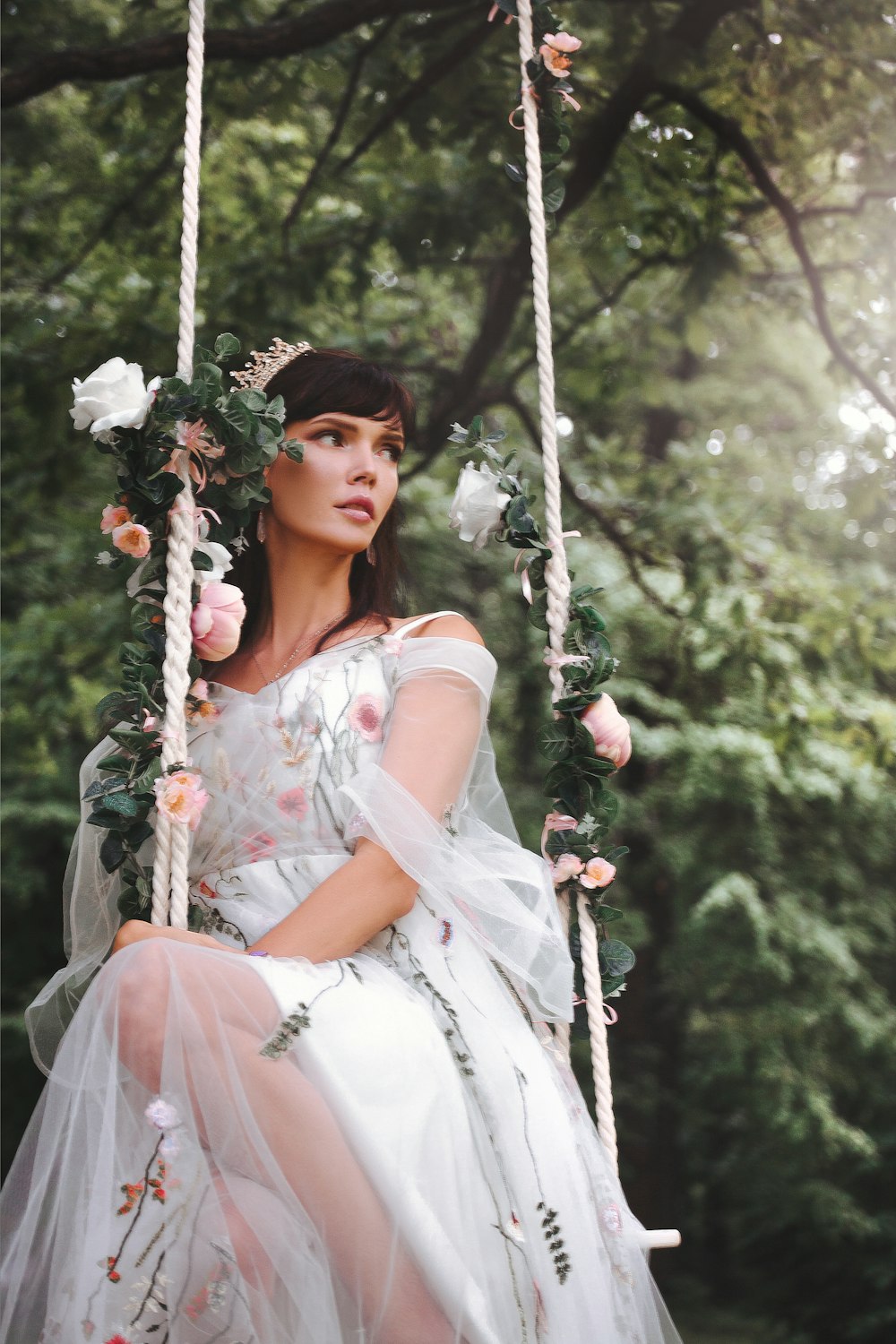 woman wearing wedding gown sitting on outdoor swing near tall trees