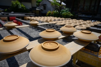 Rows of ceramic bowls placed upside down on wooden racks under the sun, likely to dry. Surrounding structures and greenery can be seen in the background, giving the scene an outdoor setting in what appears to be a pottery workshop area.