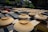 Rows of ceramic bowls placed upside down on wooden racks under the sun, likely to dry. Surrounding structures and greenery can be seen in the background, giving the scene an outdoor setting in what appears to be a pottery workshop area.