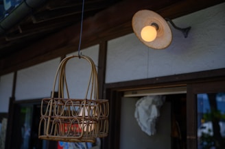 A rattan basket hangs from a wire in front of a traditional building. The building features wooden structures and a white exterior wall. Above the basket, there is an outdoor wall-mounted lamp with a warm glowing bulb. The scene appears to be taken in a rustic or vintage setting.