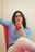 A person with long dark hair and glasses is sipping a red drink through a straw while relaxing on a cushioned seat. They are wearing a light blue sweater and white pants, and appear to be thoughtful or pensive. The background is minimal, emphasizing the relaxed and casual atmosphere.
