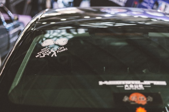 A close-up of a car rear window with a humorous sticker that features a boxing glove graphic and text that reads 'Don't touch my car' and 'Oh F##k!'. The background shows a blurred indoor setting with various vehicles partially visible.