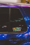 A close-up of a car window featuring a sticker with the text 'IACRO TEAM'. The car's paint appears to have an iridescent quality, reflecting various shades under the light. The window shows some reflections and blurred lights in the background.