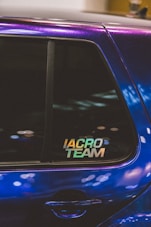 A close-up of a car window featuring a sticker with the text 'IACRO TEAM'. The car's paint appears to have an iridescent quality, reflecting various shades under the light. The window shows some reflections and blurred lights in the background.