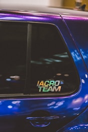 A close-up of a car window featuring a sticker with the text 'IACRO TEAM'. The car's paint appears to have an iridescent quality, reflecting various shades under the light. The window shows some reflections and blurred lights in the background.