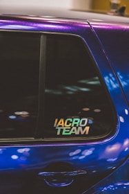 A close-up of a car window featuring a sticker with the text 'IACRO TEAM'. The car's paint appears to have an iridescent quality, reflecting various shades under the light. The window shows some reflections and blurred lights in the background.