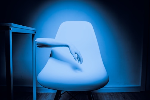 A minimalist room featuring a modern white chair in the center with a human arm extending through the backrest. The scene is lit with a blue tint, creating an ethereal and surreal atmosphere. To the left, part of a white table is visible. The wooden flooring contrasts with the sleek design of the furniture.