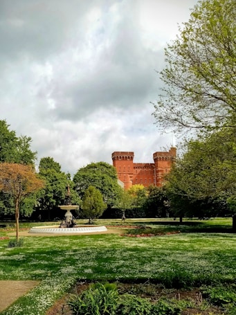 A lush park features a large, ornate fountain surrounded by a circular path and vibrant greenery. In the background, a brick castle with prominent towers rises against a cloudy sky, adding a historic and majestic feel to the scene.