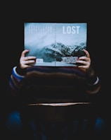 A person is sitting in a chair holding an open book in front of their face. The book's cover reads 'LOST' and features an image of mountains under a cloudy sky. The lighting is dim, creating a mysterious or contemplative atmosphere.