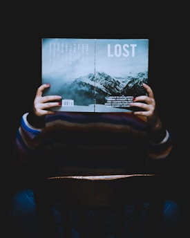 A person is sitting in a chair holding an open book in front of their face. The book's cover reads 'LOST' and features an image of mountains under a cloudy sky. The lighting is dim, creating a mysterious or contemplative atmosphere.