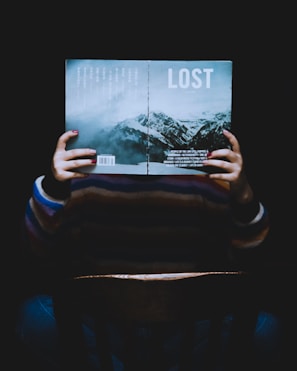 A person is sitting in a chair holding an open book in front of their face. The book's cover reads 'LOST' and features an image of mountains under a cloudy sky. The lighting is dim, creating a mysterious or contemplative atmosphere.