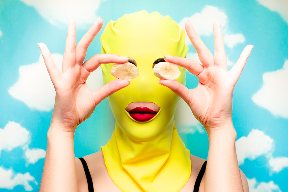 woman wearing yellow mask and covering eyes with two banana slices