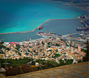 A coastal city with a busy port featuring several docked ships. The cityscape includes dense clusters of buildings with red and brown rooftops. The sea displays a gradient of blue hues, bordered by breakwaters and industrial structures, creating a harmonious blend of urban and natural elements.