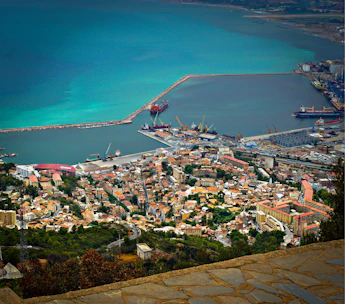 A coastal city with a busy port featuring several docked ships. The cityscape includes dense clusters of buildings with red and brown rooftops. The sea displays a gradient of blue hues, bordered by breakwaters and industrial structures, creating a harmonious blend of urban and natural elements.