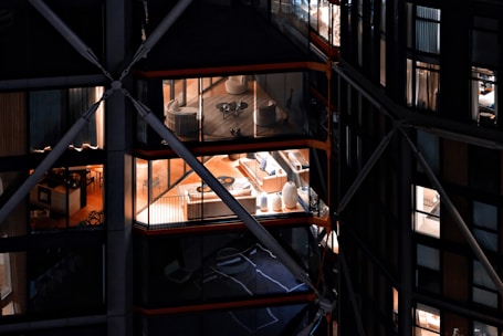 View of a modern building with large glass windows, revealing interior spaces that include elegantly furnished rooms with chairs, tables, and decorative items. The building has a distinctive architectural design with visible structural beams and crossbars.