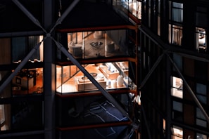 View of a modern building with large glass windows, revealing interior spaces that include elegantly furnished rooms with chairs, tables, and decorative items. The building has a distinctive architectural design with visible structural beams and crossbars.