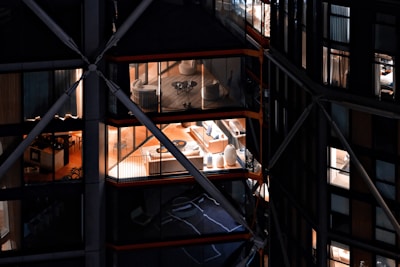 View of a modern building with large glass windows, revealing interior spaces that include elegantly furnished rooms with chairs, tables, and decorative items. The building has a distinctive architectural design with visible structural beams and crossbars.