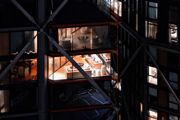 View of a modern building with large glass windows, revealing interior spaces that include elegantly furnished rooms with chairs, tables, and decorative items. The building has a distinctive architectural design with visible structural beams and crossbars.