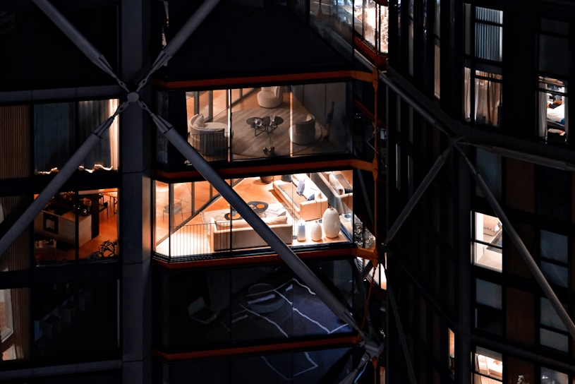 View of a modern building with large glass windows, revealing interior spaces that include elegantly furnished rooms with chairs, tables, and decorative items. The building has a distinctive architectural design with visible structural beams and crossbars.
