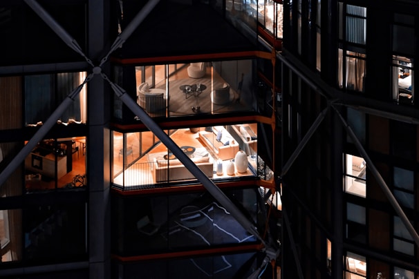 View of a modern building with large glass windows, revealing interior spaces that include elegantly furnished rooms with chairs, tables, and decorative items. The building has a distinctive architectural design with visible structural beams and crossbars.