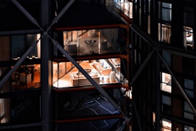 View of a modern building with large glass windows, revealing interior spaces that include elegantly furnished rooms with chairs, tables, and decorative items. The building has a distinctive architectural design with visible structural beams and crossbars.