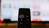 A close-up of a black remote control with visible buttons, including a prominent 'MENU' button at the top. In the background, a blurry television screen displays colorful icons and applications.