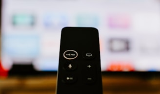 A close-up of a black remote control with visible buttons, including a prominent 'MENU' button at the top. In the background, a blurry television screen displays colorful icons and applications.