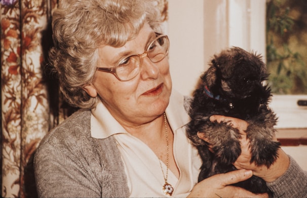 An elderly woman smiling warmly as her service dog gently rests its head on her lap at home.