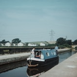 A narrowboat named 'MOHAWK' is navigating along a canal. Two people are visible on the boat, which is painted blue and features small windows and a rope at the front. The canal is bordered by a stone walkway. In the background, there are power lines and rolling green hills, as well as a row of small houses and trees.