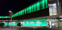 A modern airport building brightly illuminated with green lights at night. The signage reads 'Christchurch Airport' and is prominently visible. The sleek architectural design is accentuated by large glass panels and a well-lit entrance. A couple of small trees with white flowers and a red banner can be seen nearby.