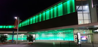 A modern airport building brightly illuminated with green lights at night. The signage reads 'Christchurch Airport' and is prominently visible. The sleek architectural design is accentuated by large glass panels and a well-lit entrance. A couple of small trees with white flowers and a red banner can be seen nearby.