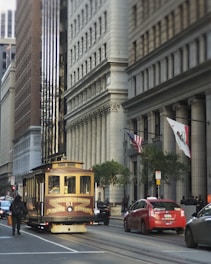 A classic streetcar marked 'Van Ness Ave. California & Market Streets' travels along a city street lined with tall buildings featuring ornate architectural details. The streetcar is surrounded by vehicles, including a red taxi, and a few pedestrians. Flags, including the American flag and the California state flag, are displayed on one of the buildings.