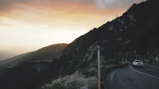 A winding mountain road with a Vibeway camper driving towards a glowing sunset.
