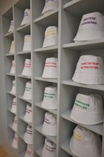 A neatly arranged display of white bucket hats sits on multiple shelves. Each hat is embroidered with words in different languages and colors, such as red, yellow, purple, and green. The hats are organized in a spacious, grid-like pattern within an indoor setting.