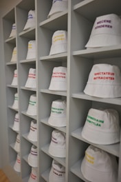 A neatly arranged display of white bucket hats sits on multiple shelves. Each hat is embroidered with words in different languages and colors, such as red, yellow, purple, and green. The hats are organized in a spacious, grid-like pattern within an indoor setting.
