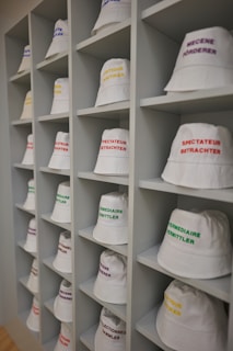 A neatly arranged display of white bucket hats sits on multiple shelves. Each hat is embroidered with words in different languages and colors, such as red, yellow, purple, and green. The hats are organized in a spacious, grid-like pattern within an indoor setting.