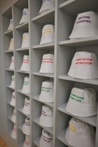 A neatly arranged display of white bucket hats sits on multiple shelves. Each hat is embroidered with words in different languages and colors, such as red, yellow, purple, and green. The hats are organized in a spacious, grid-like pattern within an indoor setting.
