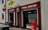 A storefront with signs for McClafferty's Bar & Lounge and Funeral Directors. The building is painted in beige with red accents. Decorative flags are displayed, and a red HB ice cream container is situated outside. A wooden bench is also visible near the entrance.