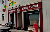 A storefront with signs for McClafferty's Bar & Lounge and Funeral Directors. The building is painted in beige with red accents. Decorative flags are displayed, and a red HB ice cream container is situated outside. A wooden bench is also visible near the entrance.