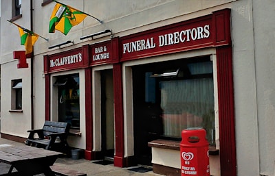 A storefront with signs for McClafferty's Bar & Lounge and Funeral Directors. The building is painted in beige with red accents. Decorative flags are displayed, and a red HB ice cream container is situated outside. A wooden bench is also visible near the entrance.